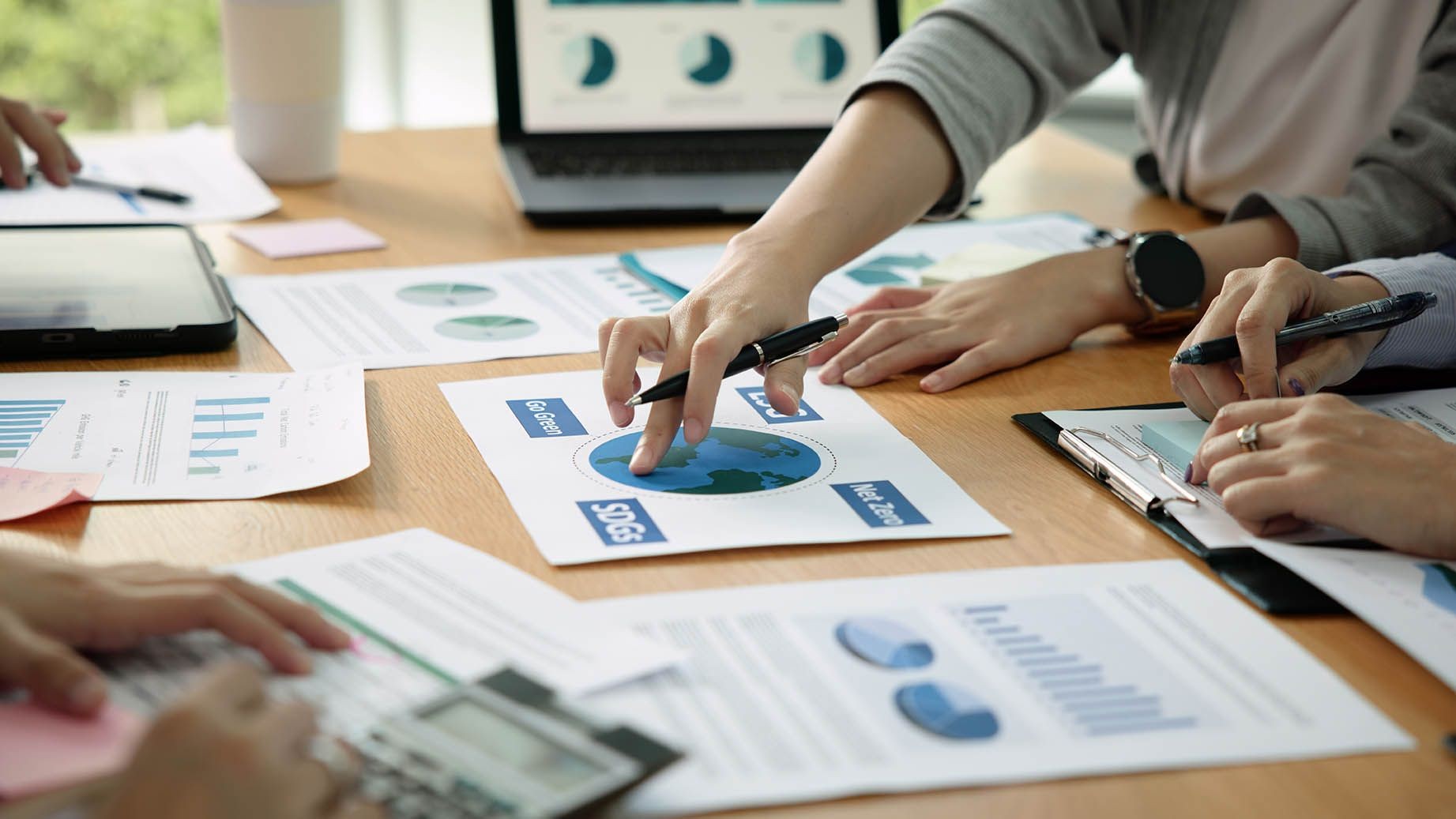 Hands holding pens above charts and reports on a desk, with a laptop in the background.