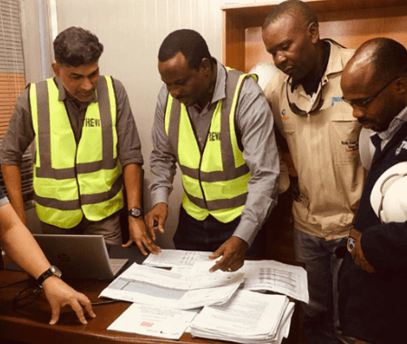 Group of four men wearing high-visibility vests reviewing documents on a table inside an office.